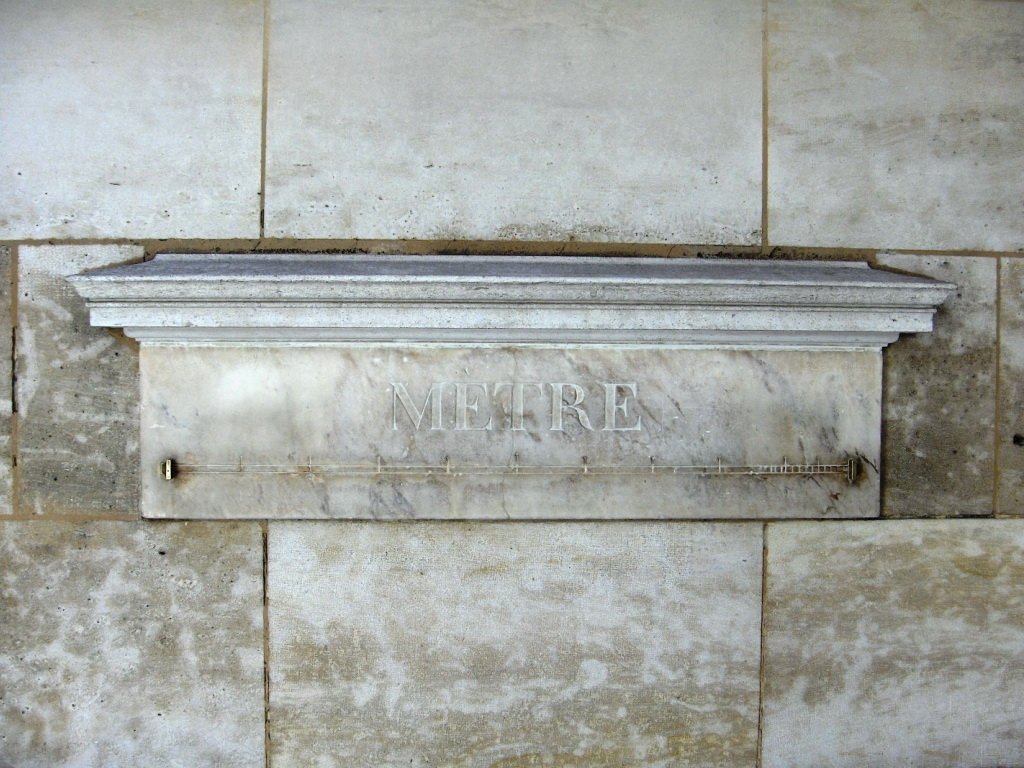A white marble plaque set into a stone wall. It is carved with the word MÈTRE, and a thin metal bar with marks runs along it between two stops.